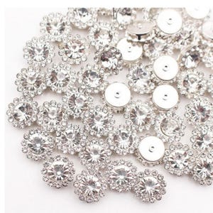 May include: A collection of silver-toned rhinestone buttons. The buttons feature a central, clear, faceted rhinestone surrounded by smaller rhinestones, creating a floral or starburst design. The backs of the buttons are silver and flat.