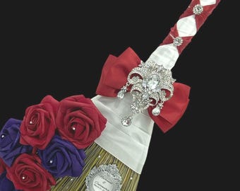 Peut inclure: Un balai décoratif avec un manche enveloppé de ruban de satin blanc, des accents de ruban rouge et des ornements en strass argentés. Une grande broche argentée à cristaux surplombe un bouquet de roses en mousse rouges et violettes et une plaque.