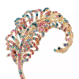 May include: A gold-toned feather-shaped brooch adorned with colorful rhinestones. The brooch features a detailed design with pink, blue, and teal stones, creating a sparkling effect. This elegant accessory is suitable for adding a touch of glamour.
