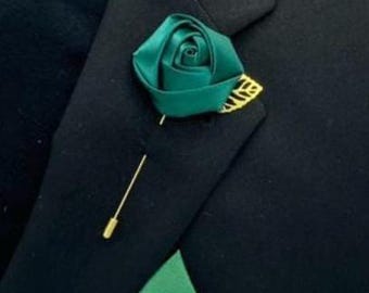 May include: A dark green satin rose lapel pin with a gold-colored pin and decorative backing. The rose is attached to a black blazer, creating a classic and elegant accessory. The pin adds a touch of sophistication to formal wear.