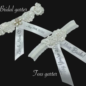 May include: Two white bridal garters. One is a traditional garter with floral lace and a pearl and rhinestone embellishment. The other is a toss garter with the text "Tag You're it!" and "Next Bride!" on the ribbon. The bridal garter has the text "Liva & Mark" and "May 18th 2026".