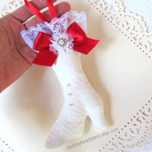May include: White fabric boot ornament with a red satin bow and white lace trim. The boot has pearl-like buttons and a small white flower with a rhinestone center.