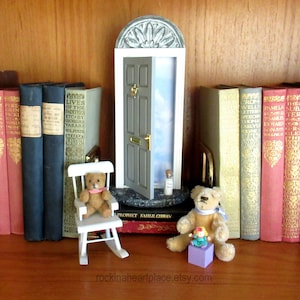 May include: A miniature gray door with an arched top, ajar, revealing a sky backdrop. A small teddy bear sits in a white rocking chair. Other teddy bears and books are arranged on a wooden surface. The Etsy shop name is visible.