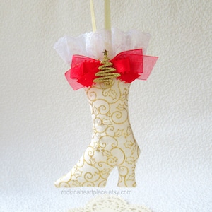 May include: A decorative Christmas ornament shaped like a high-heeled boot. The boot is cream-colored with gold holly and swirl patterns. It features a red bow, gold tree, and white lace trim. A gold ribbon is attached for hanging.