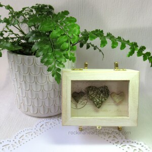 May include: Three green heart-shaped stones are displayed in a white wooden shadow box with a glass front. The box has gold hinges and a white background with a vintage design. The text "rockinaheartplace.etsy.com" is printed on the bottom of the box.