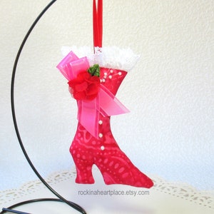 May include: A red fabric boot-shaped ornament with a high heel, adorned with a pink bow, red flower, and faux pearls. The boot has a white lace trim at the top and a red ribbon for hanging. The Etsy shop name "rockinaheartplace.etsy.com" is visible.