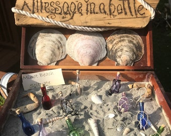 May include: A wooden box with a sign that says "Send a message in a bottle". The box is filled with sand and seashells. There are several small glass bottles with cork tops, some with colorful liquid inside. A handwritten note says "Minot Beach Scituate".