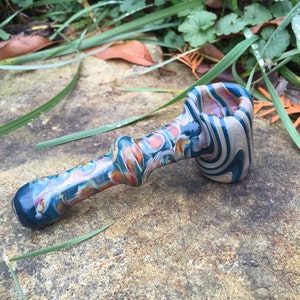 May include: A small, hand-blown glass pipe with a swirling blue, white and orange pattern. The pipe has a rounded bowl and a long, thin stem.