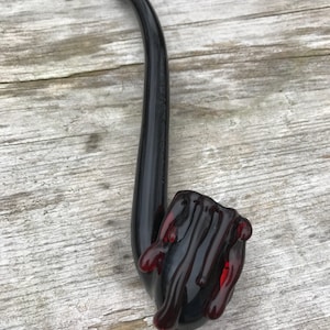 Könnte beinhalten: Eine schwarze Glaspfeife mit einem roten Glaskopf. Der Kopf ist handförmig mit tropfendem roten Glas.
