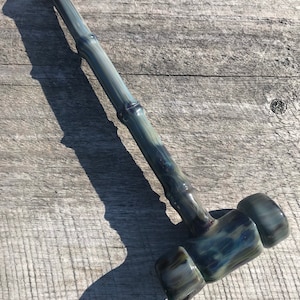 Puede incluir: Una pipa de martillo de vidrio negra y verde con un tallo largo y delgado. La pipa tiene un patrón de remolino y está hecha de vidrio grueso.