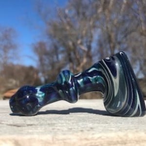 May include: A hand-blown glass pipe with a swirling pattern of dark blue, teal, and white. The pipe has a curved design with a bowl at one end and a mouthpiece at the other. The background features a blurred view of trees and a blue sky.