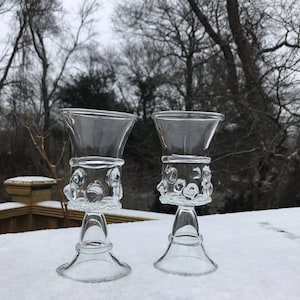 May include: Two clear glass goblets with a textured, bubble-like design on the stem and base. The goblets are sitting on a white surface.