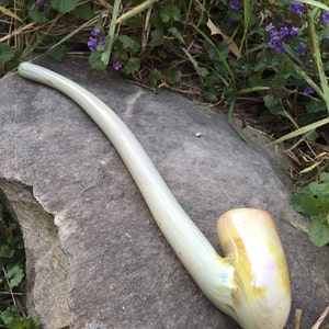 Op de afbeelding: Een witte glazen pijp met een lange, gebogen steel en een ronde kom. De pijp heeft een iriserende afwerking en ligt op een grijze rots.