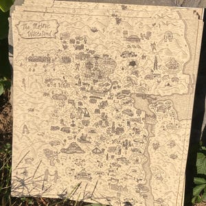 May include: A stack of beige-colored paper prints featuring a detailed, hand-drawn map. The map depicts a landscape with various structures, roads, and geographical features. The text "The Mojave Wasteland" is visible in the upper left corner. The prints are resting on a stone surface.