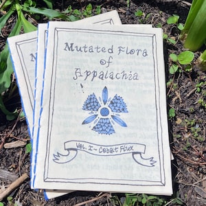 May include: A stack of three small, cream-colored books with blue stitching. The top book has the title "Mutated Flora of Appalachia" and a blue floral illustration. The text below the illustration reads "Vol. I - Cobalt Flux".