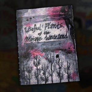 May include: A hand-painted journal titled "Useful Plants of the Mojave Wasteland." The cover features a watercolour design with black, pink, and grey hues. The title is handwritten in black ink, and there are illustrations of plants.