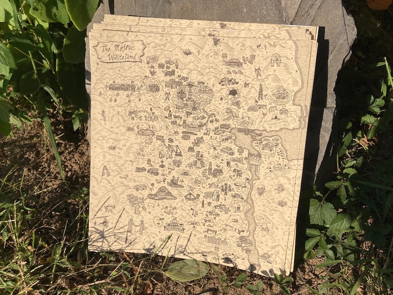May include: A stack of beige-colored paper prints featuring a detailed, hand-drawn map. The map depicts a landscape with various structures, roads, and geographical features. The text "The Mojave Wasteland" is visible in the upper left corner. The prints are resting on a stone surface.