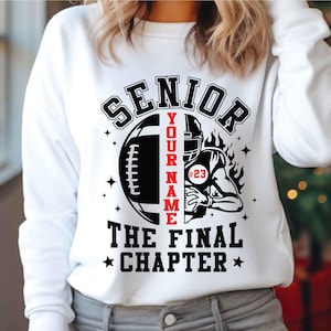 May include: White sweatshirt with a black and red football design. The design includes the word "SENIOR", a football, a football player, and the text "YOUR NAME" and "THE FINAL CHAPTER". The number "23" is on the player's jersey.