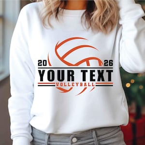Peut inclure: Sweat-shirt blanc avec un graphisme de ballon de volley-ball orange. Le design comprend le texte "YOUR TEXT" en gras noir, avec le mot "VOLLEYBALL" en dessous. Les chiffres "20" et "26" encadrent le haut du design.