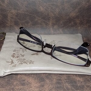 May include: A black and brown eyeglasses case with a floral pattern. The case has a zipper closure and a metal ring. A pair of black eyeglasses with clear lenses is resting on top of the case.