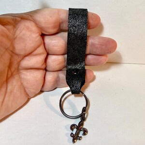 May include: Black leather keychain with a silver-colored fleur-de-lis charm. The keychain features a black metal ring and a textured leather strap. The fleur-de-lis charm adds a decorative touch to this accessory.