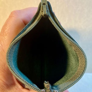 Puede incluir: Una bolsa verde con cremallera y un tirador de cremallera plateado. La bolsa est&aacute; abierta y el interior est&aacute; forrado con un tejido verde claro.