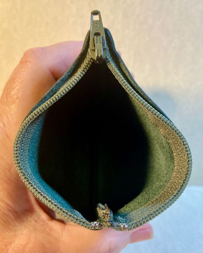 Puede incluir: Una bolsa verde con cremallera y un tirador de cremallera plateado. La bolsa est&aacute; abierta y el interior est&aacute; forrado con un tejido verde claro.