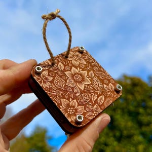 May include: A small wooden square ornament with a floral design, featuring a brown wood background and a light brown floral pattern. The ornament has a twine loop for hanging and silver metal accents.