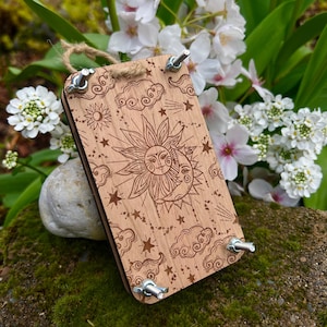 May include: A wooden sun and moon tapestry kit with a celestial design featuring stars, clouds, and shooting stars. The kit includes a wooden frame, embroidery floss, and a needle.