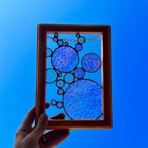 May include: A framed stained glass piece with a design of blue bubbles. The bubbles are arranged in a cluster and have a textured, iridescent finish.