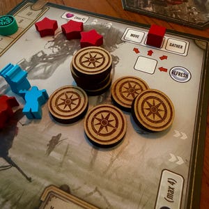 May include: A board game setup featuring a stack and several individual circular wooden tokens engraved with a compass rose design. The board includes red star-shaped pieces, a red cube, blue character figures, and text labels MOVE, GATHER, and REFRESH.