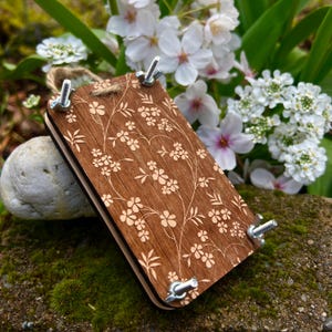 Op de afbeelding: Een houten weefgetouw met een bloemenpatroon, van lichtbruin hout met een wit bloemenpatroon. Het weefgetouw heeft vier metalen klemmen aan de zijkanten.