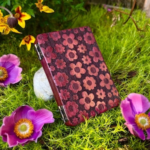 May include: A wooden box with a floral pattern in shades of red and pink, set against a backdrop of green moss and vibrant flowers. The box is rectangular with a hinged lid and metal hardware.