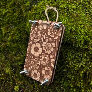 Puede incluir: Un marco de madera con estampado floral, cuatro tornillos de metal y un lazo de cuerda para colgar. El marco es marrón con un patrón floral blanco.