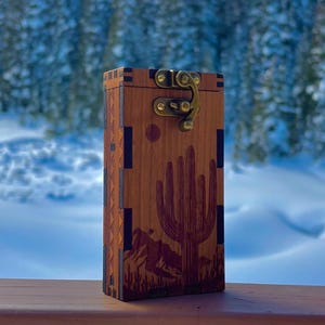 May include: A rectangular wooden box featuring a laser-engraved desert landscape with a large saguaro cactus and mountains. The box has dark wood corner accents, a geometric side pattern, and a brass-colored metal latch closure on the front.