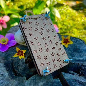 May include: A wooden flower press featuring a floral pattern, secured with four wing nuts. The press is light brown with dark brown flower designs. A natural jute cord is attached for hanging. The press is shown outdoors with flowers.