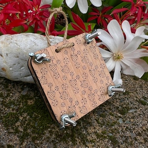 May include: A wooden flower press with a strawberry and flower pattern. The press is square, with metal wing nuts and bolts at each corner. A jute rope loop is attached for hanging. The background includes white and red flowers.