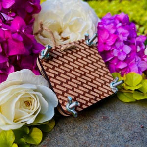 May include: A wooden flower press with a woven pattern, secured by wing nuts. The press is surrounded by vibrant purple hydrangeas, white and cream roses, and green foliage, creating a colorful floral arrangement.