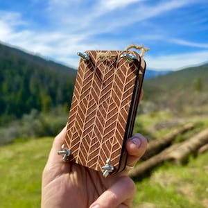 May include: A wooden plant press with a leaf pattern carved into the surface. The press is held together with four metal wing nuts and a twine loop. The background features a mountain range and blue sky.