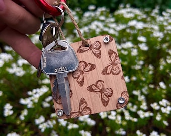 Puede incluir: Un llavero de madera con un patrón de mariposa y una llave plateada con el texto "SUZ11" en él. El llavero está unido a un mosquetón rojo.