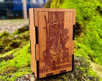 May include: A wooden box with a red-brown stained finish and black trim. The box features an engraved image of a tarot card, the Queen of Wands, with a woman sitting on a throne.