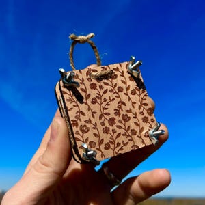 Puede incluir: Un pequeño cuadrado de madera con un patrón floral y abrazaderas de metal en cada esquina. El cuadrado se sostiene en una mano contra un cielo azul.