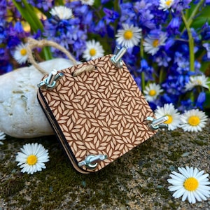May include: A small wooden square loom with a leaf pattern, four metal clamps, and a length of twine attached. The loom is sitting on a rock with blue and white flowers in the background.