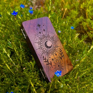 Puede incluir: Una caja rectangular de madera oscura con un diseño celestial que presenta una luna creciente, estrellas y un patrón de sol. La caja tiene una tapa con bisagras y herrajes metálicos. Rodeada de follaje verde y pequeñas flores azules.