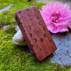Peut inclure: Une boîte rectangulaire en bois avec un motif céleste. La boîte est en bois brun rougeâtre et présente un motif gravé d'étoiles, de lunes et d'un éclat de soleil central. La boîte a des charnières et un fermoir en laiton. Une fleur rose est en arrière-plan.