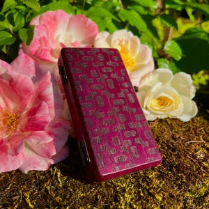 May include: A rectangular, burgundy wooden box with a geometric pattern etched into the surface. The box is hinged and has metal hardware. The background features pink and yellow roses and moss.