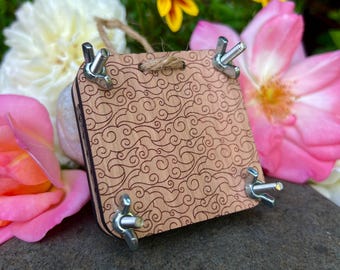 May include: A wooden flower press with a swirling pattern, secured by four wing nuts. A twine loop is attached for hanging. The press is surrounded by pink, white, and yellow flowers.