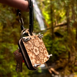 以下が含まれることがあります： 花柄のデザインと金属製のアクセントが付いた木製キーホルダー。キーホルダーは黒いカラビナに付けられています。