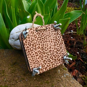 May include: A wooden plant press with a leopard print design. The press is held together with four wing nuts and bolts. A natural rope loop is attached for hanging. The press is displayed outdoors with greenery in the background.
