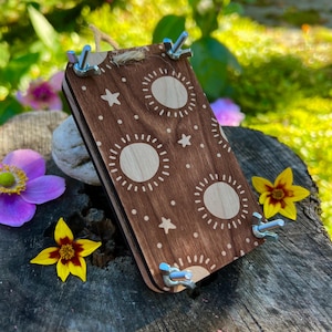 Può includere: Una pressa per fiori in legno con un motivo decorativo a sole e stelle. La pressa è marrone con disegni di sole chiari e punti bianchi. Ha dadi ad alette in metallo e un anello di spago per appenderla. Fiori gialli e viola circondano la pressa.